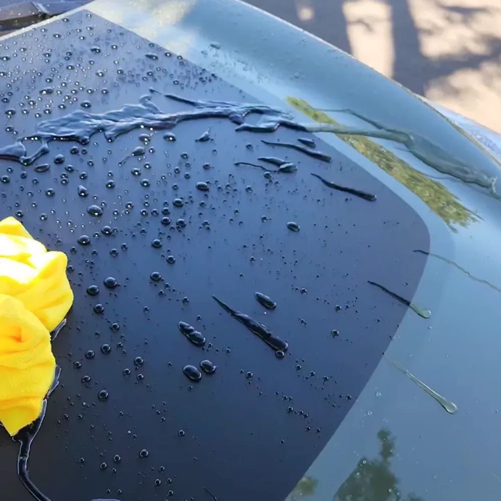 Water beading tightly on the ceramic coated hood of a dark car, with a yellow microfiber towel at the edge of the frame